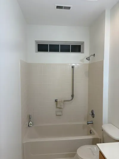 Dated beige tiled tub and shower area in a bathroom before renovation, with the toilet still wrapped in plastic.