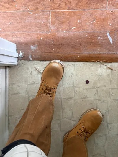 Worker's boots stand at the transition between wood-look laminate and patterned vinyl tile flooring.