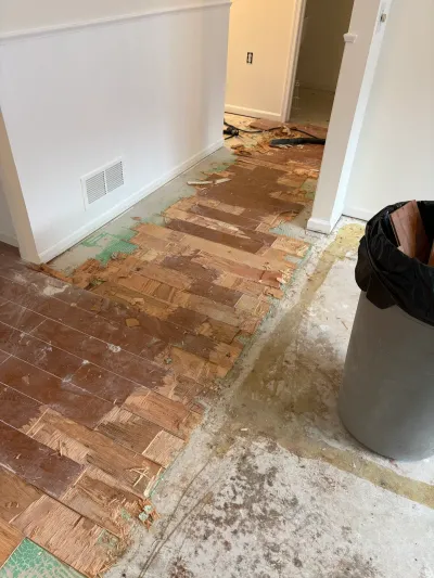 Partially demolished hardwood floor with exposed subfloor, debris, and a trash can of discarded flooring.