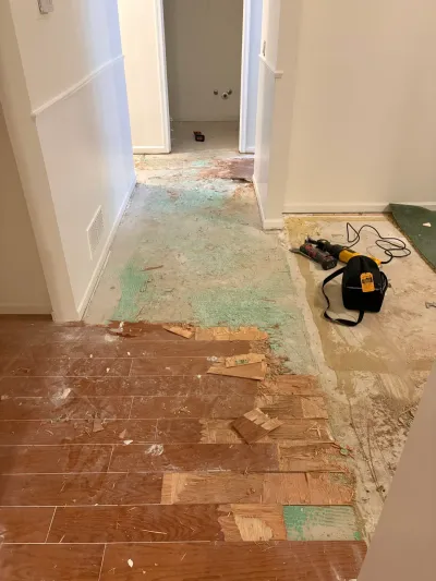 Hallway mid-renovation with flooring torn up, exposing subfloor, adhesive residue, and power tools.