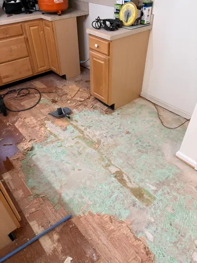 Kitchen mid-renovation with parquet flooring partially removed, exposing plywood subfloor and adhesive residue.