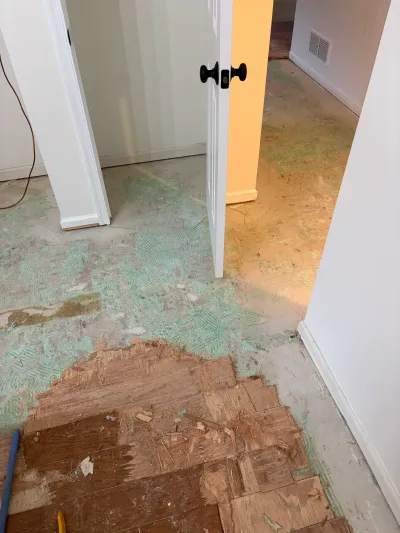 A hallway mid-renovation with exposed subfloor, adhesive residue, and a partially removed plywood floor section.