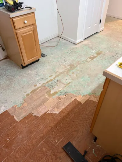 Kitchen floor mid-demolition with vinyl plank flooring partly removed, exposing green adhesive on the subfloor.