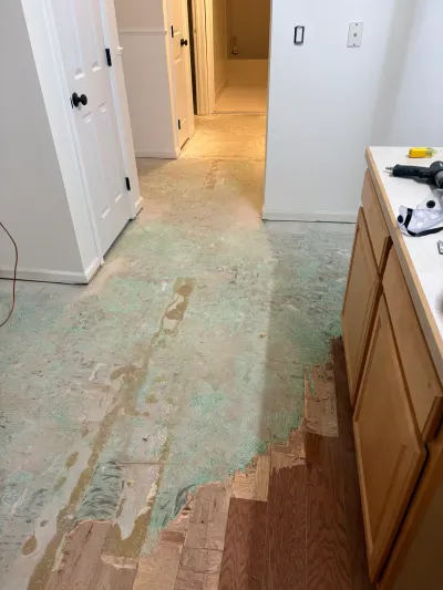 Hallway mid-renovation with old flooring removed, exposing a subfloor with adhesive residue near a cabinet.