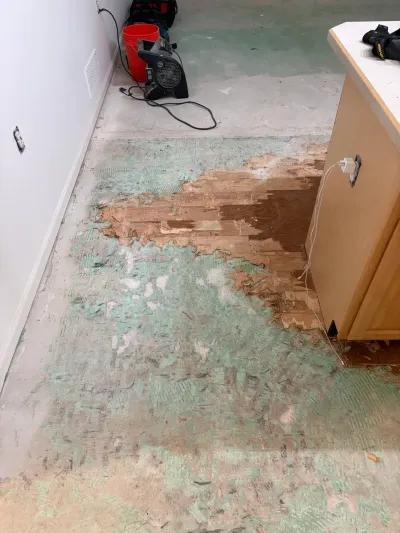 Kitchen floor mid-demolition with old flooring peeled back to concrete and a drying fan running nearby.