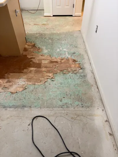 Kitchen subfloor mid-demolition showing exposed concrete, adhesive residue, and remnants of old parquet flooring.