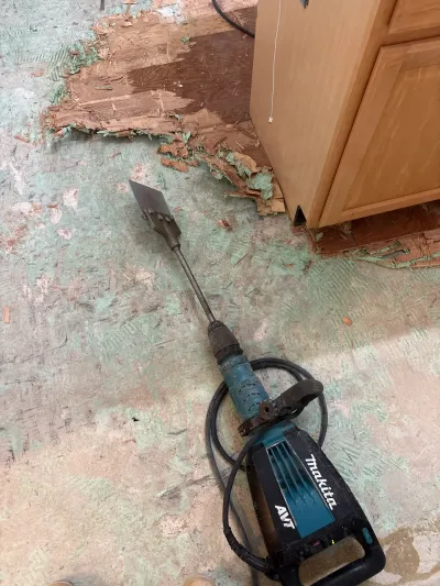 A demolition scraper tool lies on a kitchen subfloor being stripped of old flooring near wood cabinets.