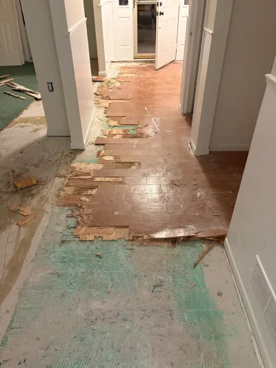 Hallway mid-renovation with wood flooring partially removed, exposing concrete subfloor and adhesive residue.