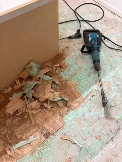 A demolition hammer sits on a subfloor covered in torn-up flooring debris and green adhesive residue.