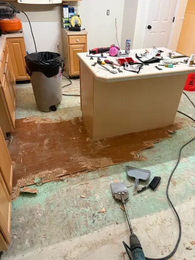 Kitchen mid-renovation with old flooring stripped away, exposing subfloor and layers of underlayment near an island.
