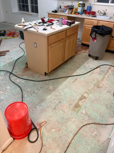 A kitchen mid-renovation with subfloor exposed, tools scattered on an island, and cords running across the floor.