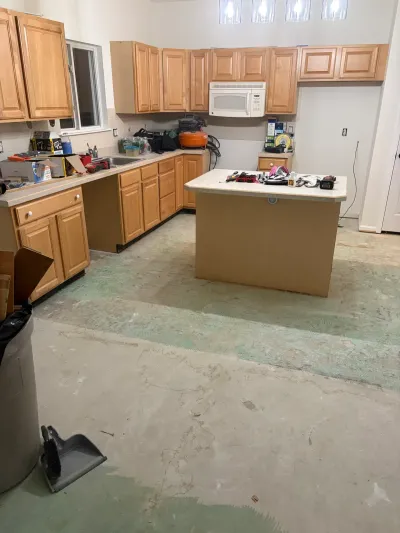 Kitchen mid-renovation with oak cabinets intact but flooring removed down to bare concrete.