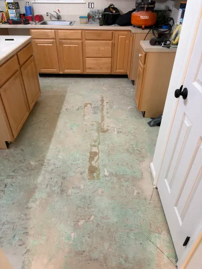 Kitchen mid-renovation with flooring stripped to the subfloor and tools staged on the counters.