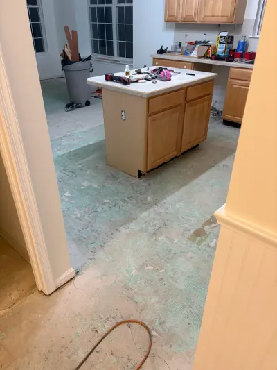 A kitchen mid-renovation with flooring removed, exposed adhesive residue on the subfloor, and tools scattered on the island.