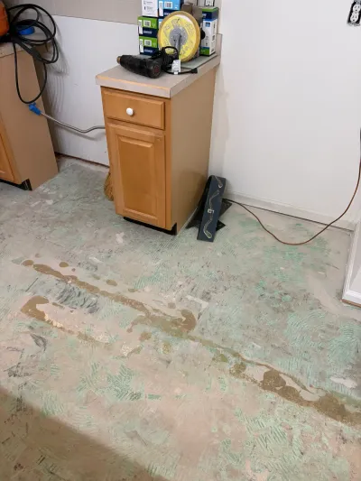 Kitchen mid-renovation with bare concrete subfloor, adhesive residue, and a single cabinet remaining.