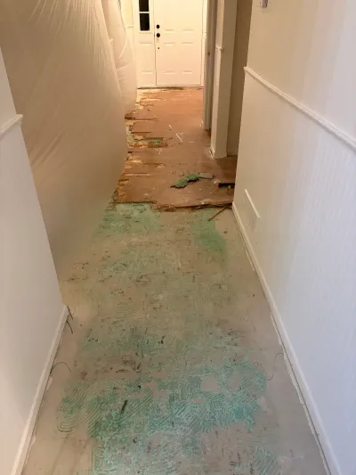 Hallway with flooring removed down to subfloor and adhesive residue, plastic sheeting protecting the wall, leading to a front