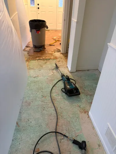 A hallway mid-demolition with subfloor exposed, adhesive residue, and a floor scraper tool on the ground.