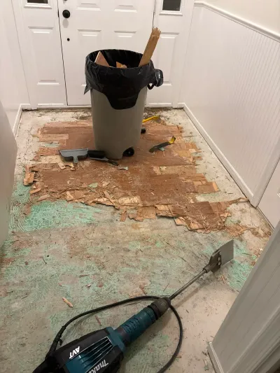 Entryway floor mid-demolition with old flooring scraped away and a scraper tool in the foreground.