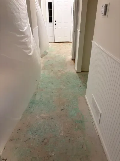 Hallway subfloor exposed during flooring removal, with plastic sheeting protecting the walls near the front door.