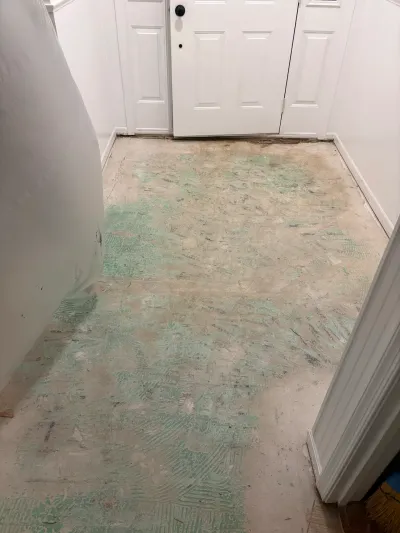 Entryway subfloor exposed during demolition, with green adhesive residue and a white front door ahead.
