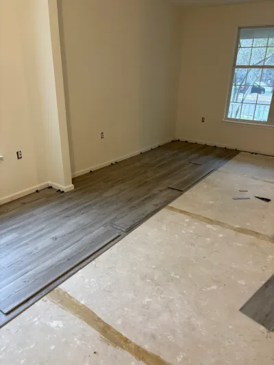 A living room mid-renovation with new gray plank flooring partially installed over an exposed plywood subfloor.