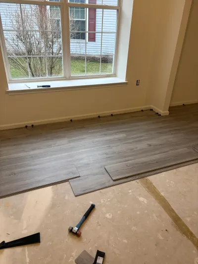 Gray vinyl plank flooring being installed over a bare subfloor in a sunlit living room.