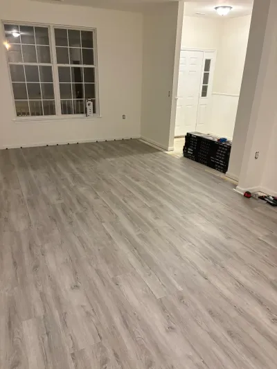A living room mid-renovation with new gray wood-look vinyl plank flooring and flooring boxes stacked near the entry.