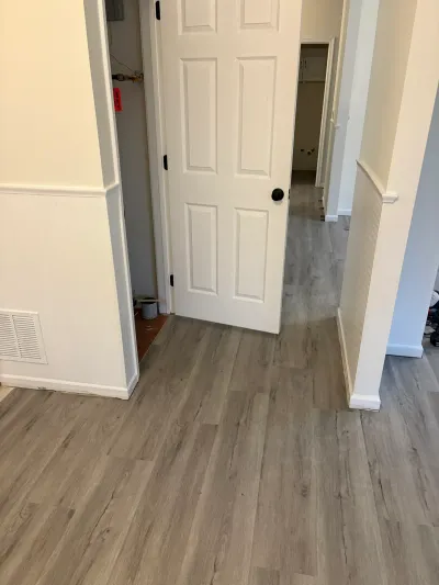 A hallway with new gray vinyl plank flooring, an open door revealing a utility closet with exposed plumbing.