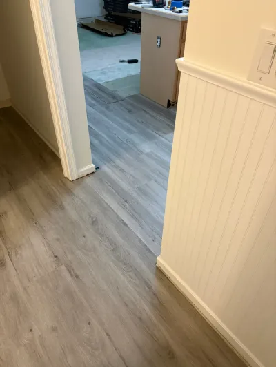 A hallway with new gray plank flooring leads into a kitchen area mid-renovation with flooring materials on the floor.
