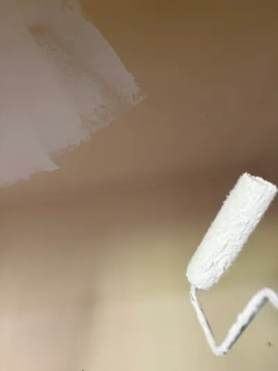 Close-up of a paint roller applying white paint over a tan wall or ceiling surface.