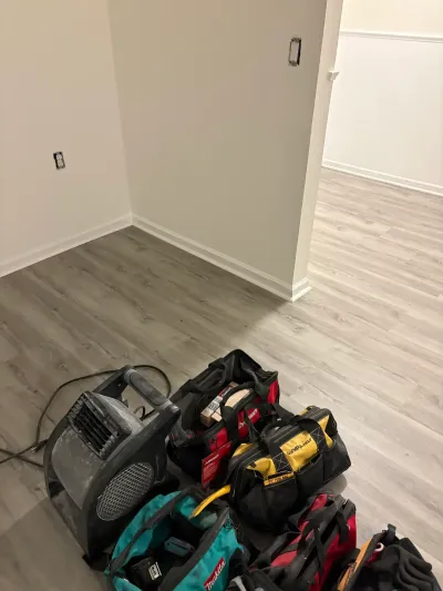 Basement under renovation with new gray vinyl plank flooring, tool bags, and an air mover fan.
