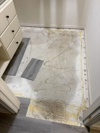 Subfloor with adhesive residue exposed during flooring removal, with new gray plank samples laid nearby.