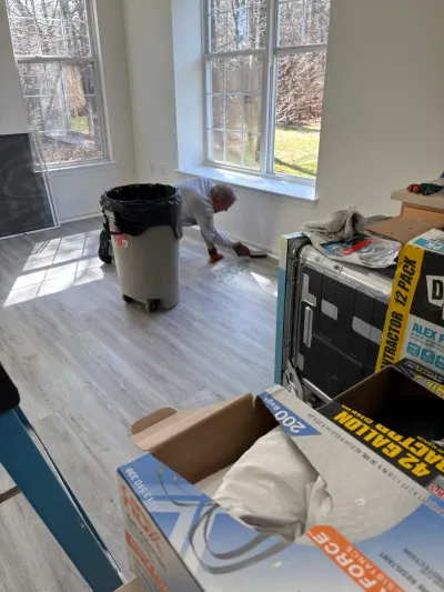 A worker finishes new plank flooring in a room filled with renovation supplies and windows.