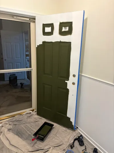 A door leans against a wall, partially painted olive green, with painting supplies on a drop cloth below.