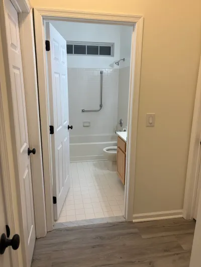 A finished bathroom with tiled tub-shower, oak vanity, and tile flooring, seen through an open hallway door.