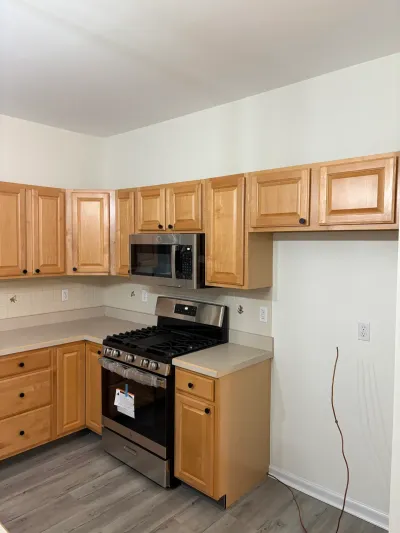 A kitchen mid-renovation with oak cabinets, a new range still wrapped in plastic, and an unfinished wall with exposed wiring.