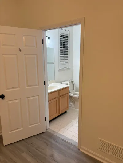 View through an open door into a small bathroom with oak vanity, tile floor, and shuttered window before renovation.