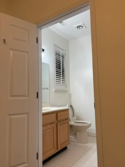 A small bathroom with an oak vanity, toilet, and shuttered window, seen through an open door.