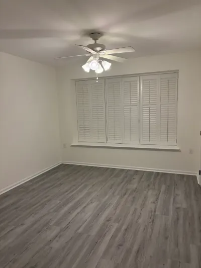 Empty renovated room with gray plank flooring, white plantation shutters, and a lit ceiling fan.