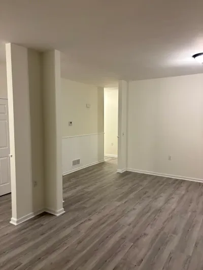 Empty finished basement room with gray vinyl plank flooring and white walls leading to a hallway.
