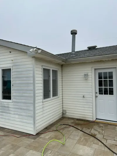 Corner of a house exterior with vinyl siding, windows, and a white door above a stone patio on an overcast day.