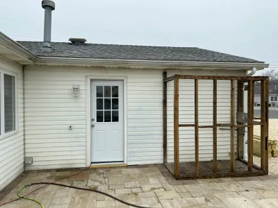 Side of a house with a new door installed next to an unfinished wood-framed addition on a paver patio.