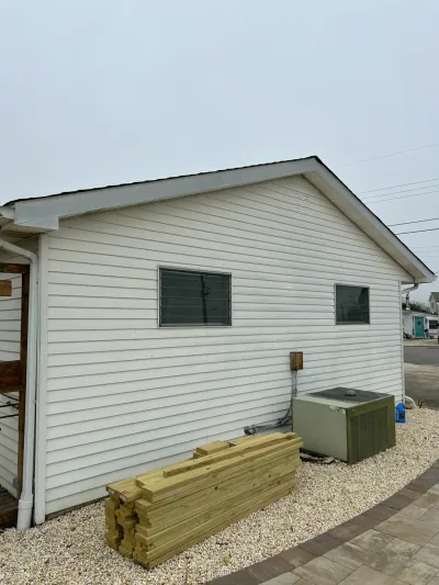 Side of a house with white vinyl siding, an HVAC unit, and stacked lumber on a gravel bed.