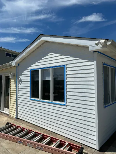 A house gable wall with new white vinyl siding partially installed, windows taped off, and a ladder on the patio below.