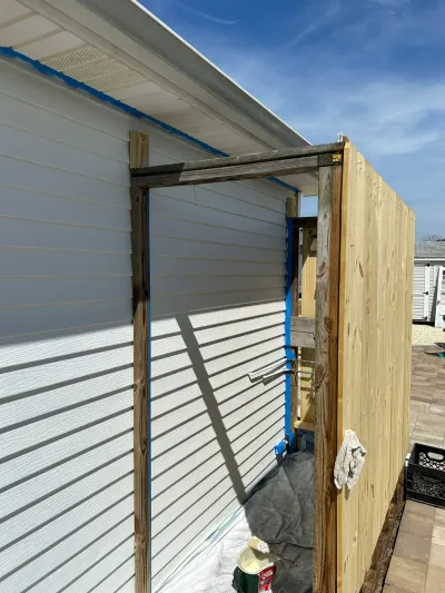 A wood enclosure under construction is being attached to the corner of a house with vinyl siding on a sunny day.