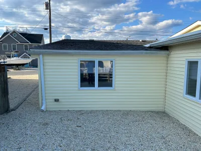 Side view of a house with yellow vinyl siding, white-trimmed windows, and a gravel yard under a partly cloudy sky.