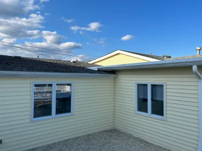Exterior corner of a house with yellow siding, blue-trimmed windows, and a gravel yard under a blue sky.