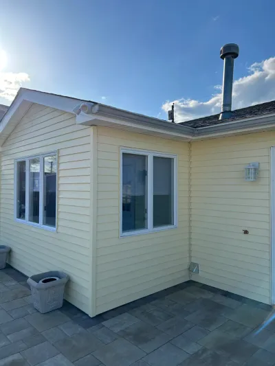 Corner of a house with fresh pale yellow siding, white-trimmed windows, and a paved patio in front.
