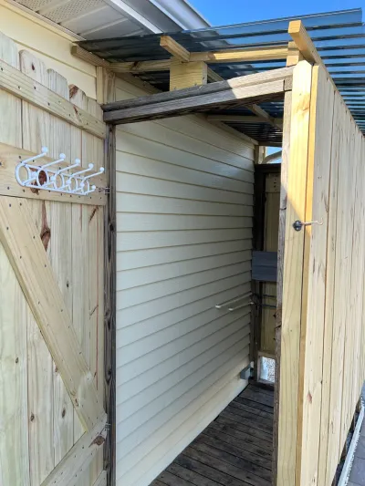 Open wooden outdoor shower enclosure with a hook rack on the door and vinyl siding visible inside.