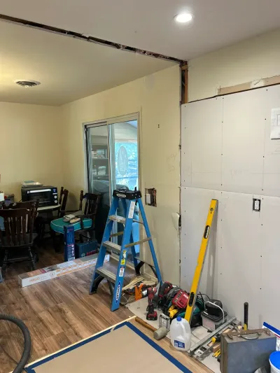 Kitchen renovation in progress with new drywall, a ladder, tools, and boxed flooring beside a dining area.